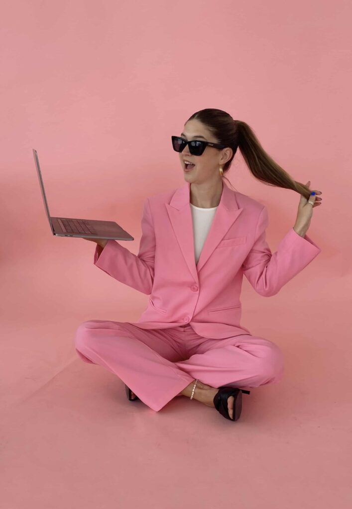 Woman wearing pink blazer set sitting on ground with laptop